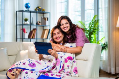 Tablet bilgisayar kullanan yaşlı bir anne, sari giymiş Hintli yaşlı bir anne kanepenin arkasından genç kızı kucaklarken hem kaydırıyor, hem sörf yapıyor, hem de modern iç mekanlarda keyif veren dijital cihazlardan alışveriş yapıyor.