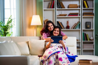Tablet bilgisayar kullanan yaşlı bir anne, sari giymiş Hintli yaşlı bir anne kanepenin arkasından genç kızı kucaklarken hem kaydırıyor, hem sörf yapıyor, hem de modern iç mekanlarda keyif veren dijital cihazlardan alışveriş yapıyor.