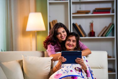 Tablet bilgisayar kullanan yaşlı bir anne, sari giymiş Hintli yaşlı bir anne kanepenin arkasından genç kızı kucaklarken hem kaydırıyor, hem sörf yapıyor, hem de modern iç mekanlarda keyif veren dijital cihazlardan alışveriş yapıyor.