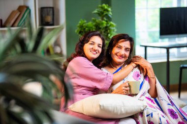 Yaşlı anne kız sohbet ediyor. Kanepede otururken el ele tutuşuyor. Hintli Asyalı yaşlı anne sari içinde ve genç kız yerde oturuyor.