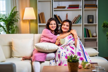 Yaşlı anne kız sohbet ediyor. Kanepede otururken el ele tutuşuyor. Hintli Asyalı yaşlı anne sari içinde ve genç kız yerde oturuyor.