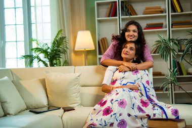 Hintli kıdemli anne kızı koltukta sarılıyor. Asyalı genç kız yaşlı anneye sürpriz yapıyor. Kitap okuyor, hem sohbet ediyor hem de modern oturma odasında mutlu bir bağ kuruyor.