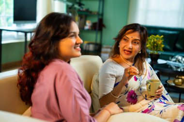 Anne kız kahve içerken sohbet ediyor, Hintli yaşlı anne sari içinde genç kızıyla birlikte modern oturma odasında koltukta oturuyor, hayat deneyimlerini paylaşıyor, birlikte sıcak kahve içerken gülümsüyor.