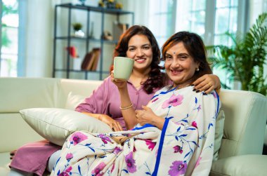 Anne kız kahve içerken sohbet ediyor, Hintli yaşlı anne sari içinde genç kızıyla birlikte modern oturma odasında koltukta oturuyor, hayat deneyimlerini paylaşıyor, birlikte sıcak kahve içerken gülümsüyor.