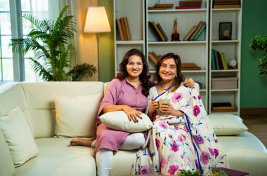 Anne kız kahve içerken sohbet ediyor, Hintli yaşlı anne sari içinde genç kızıyla birlikte modern oturma odasında koltukta oturuyor, hayat deneyimlerini paylaşıyor, birlikte sıcak kahve içerken gülümsüyor.