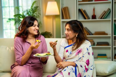 Büyük anne kız mango yiyor, Hintli yaşlı anne sari içinde genç kız kanepede oturuyor rahat modern evde taze yeşil mango dilimlerinin tadını çıkarıyor neşeli anı paylaşıyorlar.