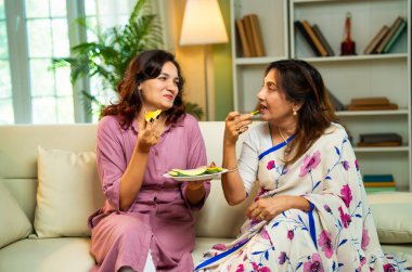 Büyük anne kız mango yiyor, Hintli yaşlı anne sari içinde genç kız kanepede oturuyor rahat modern evde taze yeşil mango dilimlerinin tadını çıkarıyor neşeli anı paylaşıyorlar.