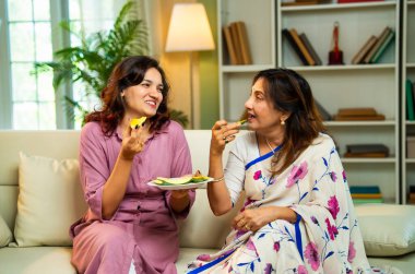 Büyük anne kız mango yiyor, Hintli yaşlı anne sari içinde genç kız kanepede oturuyor rahat modern evde taze yeşil mango dilimlerinin tadını çıkarıyor neşeli anı paylaşıyorlar.