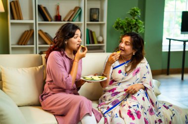 Büyük anne kız mango yiyor, Hintli yaşlı anne sari içinde genç kız kanepede oturuyor rahat modern evde taze yeşil mango dilimlerinin tadını çıkarıyor neşeli anı paylaşıyorlar.