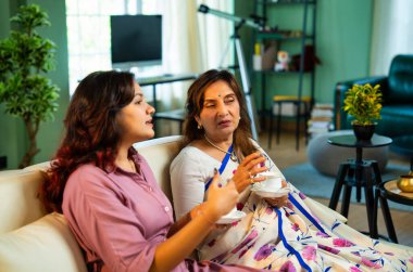 Büyük anne kız çay içiyor, Hintli yaşlı anne sari içinde genç kızıyla birlikte modern oturma odasındaki kanepede oturuyor sıcak sabah içeceğinin tadını çıkarıyor neşeyle sohbet ederken