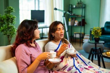 Büyük anne kız çay içiyor, Hintli yaşlı anne sari içinde genç kızıyla birlikte modern oturma odasındaki kanepede oturuyor sıcak sabah içeceğinin tadını çıkarıyor neşeyle sohbet ederken