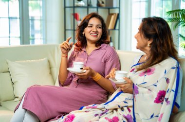 Büyük anne kız çay içiyor, Hintli yaşlı anne sari içinde genç kızıyla birlikte modern oturma odasındaki kanepede oturuyor sıcak sabah içeceğinin tadını çıkarıyor neşeyle sohbet ederken