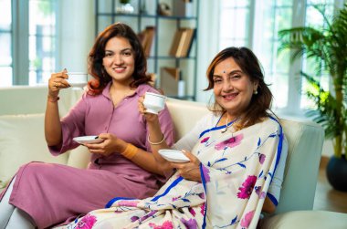 Büyük anne kız çay içiyor, Hintli yaşlı anne sari içinde genç kızıyla birlikte modern oturma odasındaki kanepede oturuyor sıcak sabah içeceğinin tadını çıkarıyor neşeyle sohbet ederken