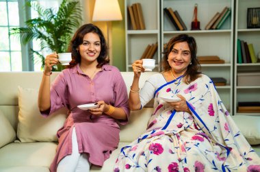 Büyük anne kız çay içiyor, Hintli yaşlı anne sari içinde genç kızıyla birlikte modern oturma odasındaki kanepede oturuyor sıcak sabah içeceğinin tadını çıkarıyor neşeyle sohbet ederken