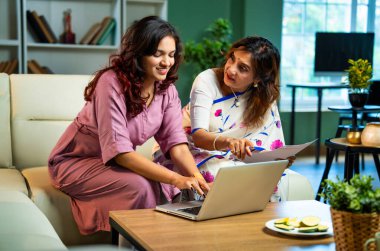 Laptop ve belgelerle muhasebe yapan bir anne, sari içinde Hintli yaşlı bir anne ve modern oturma odasındaki koltukta oturan genç bir kız, raporlar ve bilgisayar kullanarak ev ekonomisini tartışıyorlar.