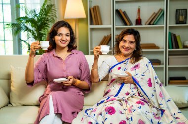 Büyük anne kız çay içiyor, Hintli yaşlı anne sari içinde genç kızıyla birlikte modern oturma odasındaki kanepede oturuyor sıcak sabah içeceğinin tadını çıkarıyor neşeyle sohbet ederken