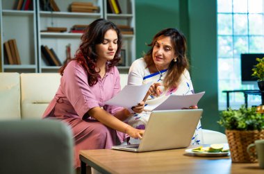 Laptop ve belgelerle muhasebe yapan bir anne, sari içinde Hintli yaşlı bir anne ve modern oturma odasındaki koltukta oturan genç bir kız, raporlar ve bilgisayar kullanarak ev ekonomisini tartışıyorlar.