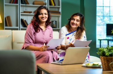 Laptop ve belgelerle muhasebe yapan bir anne, sari içinde Hintli yaşlı bir anne ve modern oturma odasındaki koltukta oturan genç bir kız, raporlar ve bilgisayar kullanarak ev ekonomisini tartışıyorlar.