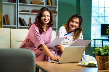 Laptop ve belgelerle muhasebe yapan bir anne, sari içinde Hintli yaşlı bir anne ve modern oturma odasındaki koltukta oturan genç bir kız, raporlar ve bilgisayar kullanarak ev ekonomisini tartışıyorlar.