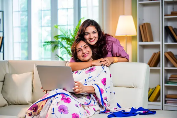 Yaşlı anne kız dizüstü bilgisayar kullanıyor, Hintli son sınıf öğrencisi sari giymiş genç kızı hem alışveriş yaparken, hem sörf yaparken, hem de modern evde kaliteli zaman geçirirken ekranda sarılıyor.