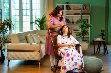 Sari içinde sallanan sandalyede oturan Hintli yaşlı bir kadına göz damlası veren ve göz yorgunluğu olan akıllı telefon kullanan kız, ev içinde göz sağlığı için sıvı ilaç uygulayarak ona yardım ediyor.