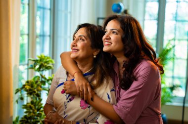 Anne kız sabah kahvesini pencerenin yanında içiyor, Hint Asyalı yaşlı anne sari içinde ve genç kız modern evde dikiliyor sıcak içkinin tadını çıkarırken doğa manzarasını izliyor ve mutlu mesut kaynaşıyor.