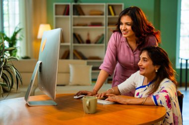 Yaşlı anne evde bilgisayar kullanıyor, Hintli Asya 'lı kıdemli anne genç kızıyla birlikte masada oturuyor masaüstü bilgisayarını kullanarak çevrimiçi sörf yapıyor, alışveriş yapıyor, işaret ediyor, kapalı mekanda mutlu bir şekilde bağ kuruyor.