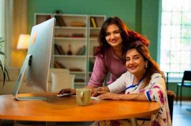 Yaşlı anne evde bilgisayar kullanıyor, Hintli Asya 'lı kıdemli anne genç kızıyla birlikte masada oturuyor masaüstü bilgisayarını kullanarak çevrimiçi sörf yapıyor, alışveriş yapıyor, işaret ediyor, kapalı mekanda mutlu bir şekilde bağ kuruyor.