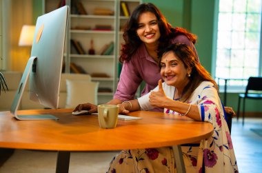 Yaşlı anne evde bilgisayar kullanıyor, Hintli Asya 'lı kıdemli anne genç kızıyla birlikte masada oturuyor masaüstü bilgisayarını kullanarak çevrimiçi sörf yapıyor, alışveriş yapıyor, işaret ediyor, kapalı mekanda mutlu bir şekilde bağ kuruyor.