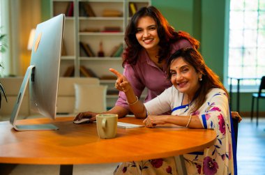 Yaşlı anne evde bilgisayar kullanıyor, Hintli Asya 'lı kıdemli anne genç kızıyla birlikte masada oturuyor masaüstü bilgisayarını kullanarak çevrimiçi sörf yapıyor, alışveriş yapıyor, işaret ediyor, kapalı mekanda mutlu bir şekilde bağ kuruyor.