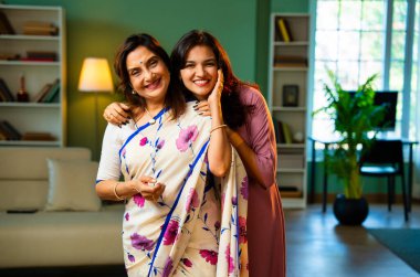 Hint kıdemli anne kızı kameraya gülümsüyor, Hintli Asyalı yaşlı anne sari içinde, genç kızı modern oturma odasında yan yana duruyor, neşe dolu yüz ifadeleriyle doğrudan ona bakıyor.