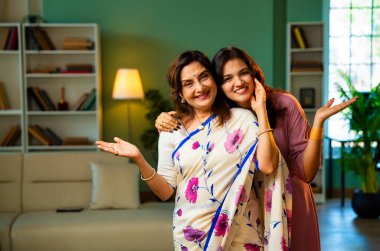 Hint kıdemli anne kızı kameraya gülümsüyor, Hintli Asyalı yaşlı anne sari içinde, genç kızı modern oturma odasında yan yana duruyor, neşe dolu yüz ifadeleriyle doğrudan ona bakıyor.