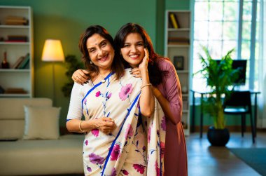 Hint kıdemli anne kızı kameraya gülümsüyor, Hintli Asyalı yaşlı anne sari içinde, genç kızı modern oturma odasında yan yana duruyor, neşe dolu yüz ifadeleriyle doğrudan ona bakıyor.