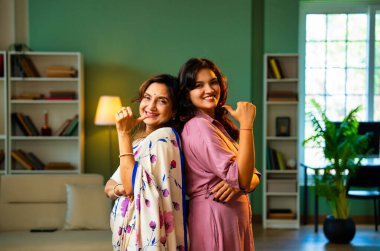 Hint kıdemli anne kızı kameraya gülümsüyor, Hintli Asyalı yaşlı anne sari içinde, genç kızı modern oturma odasında yan yana duruyor, neşe dolu yüz ifadeleriyle doğrudan ona bakıyor.