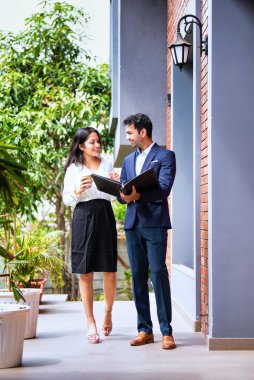 Hintli iş adamları dışarıda yürüyor, şirket adamı ve kadın modern ofis verandasında yürürken iş tartışıyorlar, dosya ve kahve tutuyorlar, takım çalışması ve profesyonel işbirliği sergiliyorlar.