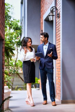 Hintli iş adamları dışarıda yürüyor, şirket adamı ve kadın modern ofis verandasında birlikte yürürken iş tartışıyorlar, tablet ve kahve tutuyorlar, takım çalışması ve işbirliği sergiliyorlar.