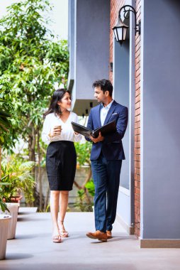 Hintli iş adamları dışarıda yürüyor, şirket adamı ve kadın modern ofis verandasında yürürken iş tartışıyorlar, dosya ve kahve tutuyorlar, takım çalışması ve profesyonel işbirliği sergiliyorlar.