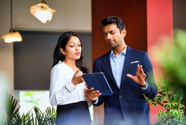 Hintli iş adamları tablet kovalamaca sürelerini kullanırken, Asyalı kurumsal erkek ve kadın profesyonel resmi giyinmiş modern ofiste dikiliyor ve tablet kullanarak proje ilerlemesini acilen tartışıyorlar.