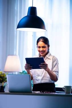 Masada oturmuş gülümseyen iş kadını neşeli bir ifadeyle kameraya bakarken, Hintli Asyalı kadın çalışan, modern samimi şirket ortamında ofis masasında dizüstü bilgisayarla güvenle poz veriyor.