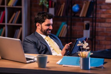 İş adamı masadaki bacakları ile tablet bilgisayarı kullanıyor, Hintli Asyalı şirket yöneticisi dijital aranın keyfini çıkarıyor, güven sembolü, teknoloji yaşam tarzı, özgürlük ve modern ofis kültürü.