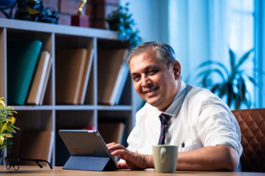 Orta çağ Hindistanlı işadamı masada otururken tablet bilgisayar kullanıyor, Asyalı şirket yöneticisi modern ofis ortamında teknolojik profesyonelliğe odaklanmış dijital cihazlar üzerinde çalışıyor.