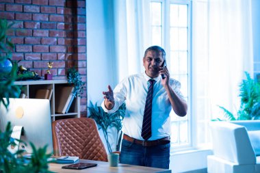 Hintli CEO kahveyle yürüyor ve modern ofiste telefon görüşmelerine katılıyor, Asyalı şirket yöneticisi kendine güvenle birden fazla iş yapıyor, iş saatlerinde şık iş yerlerinde profesyonellik yapıyor.