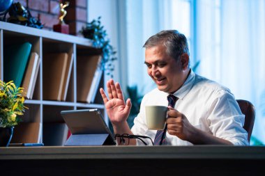 Hintli işadamı ofis masasında otururken tablet bilgisayar kullanarak video görüşmesine katılıyor, Asyalı şirket yöneticisi modern çalışma alanına güvenle sanal toplantıya katılıyor