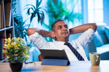 Hintli işadamı modern işyerindeki ofis masasında dinleniyor, Asyalı şirket yöneticisi sakin bir ifadeyle geriye yaslanıyor iş günü kısa bir mola veriyor, stres atmanın sembolü.