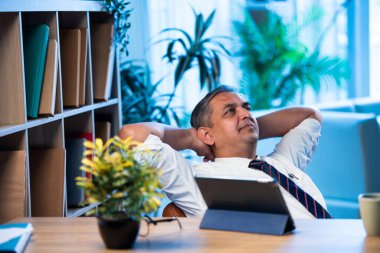 Hintli işadamı modern işyerindeki ofis masasında dinleniyor, Asyalı şirket yöneticisi sakin bir ifadeyle geriye yaslanıyor iş günü kısa bir mola veriyor, stres atmanın sembolü.