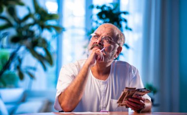 Hindistanlı kıdemli bir adam, evdeki harcamaları sayıyor, mutlu yaşlı bir insan, aylık bütçesini tatmin edici bir şekilde yönetiyor, ev ortamında rahat bir şekilde tasarruf hesaplıyor.