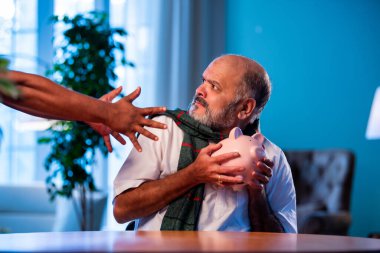 Hindistanlı kıdemli adam domuz kumbarasını hayali hırsızdan korurken modern evdeki masada oturuyor, komik bir şekilde yaşlı finansal güvenlik anlayışı, mutlu Asyalı adam içeride birikimlerini savunuyor.