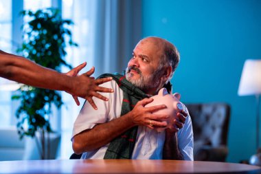 Hindistanlı kıdemli adam domuz kumbarasını hayali hırsızdan korurken modern evdeki masada oturuyor, komik bir şekilde yaşlı finansal güvenlik anlayışı, mutlu Asyalı adam içeride birikimlerini savunuyor.