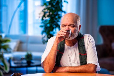 Hintli kıdemli bir adam modern evdeki masada otururken camdan su içiyor. Sağlıklı, mutlu, yaşlı, Asyalı bir erkek gündüzleri rahat bir şekilde evde susuz kalıyor.