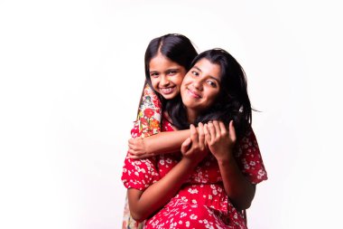 Beyaz arka planda dururken kameraya sarılıp gülümseyen Hintli küçük kardeş kızlar. Renkli elbiseler içinde Asyalı küçük kız kardeşler neşeli ifadelerle sevgi ve neşe gösteriyorlar.